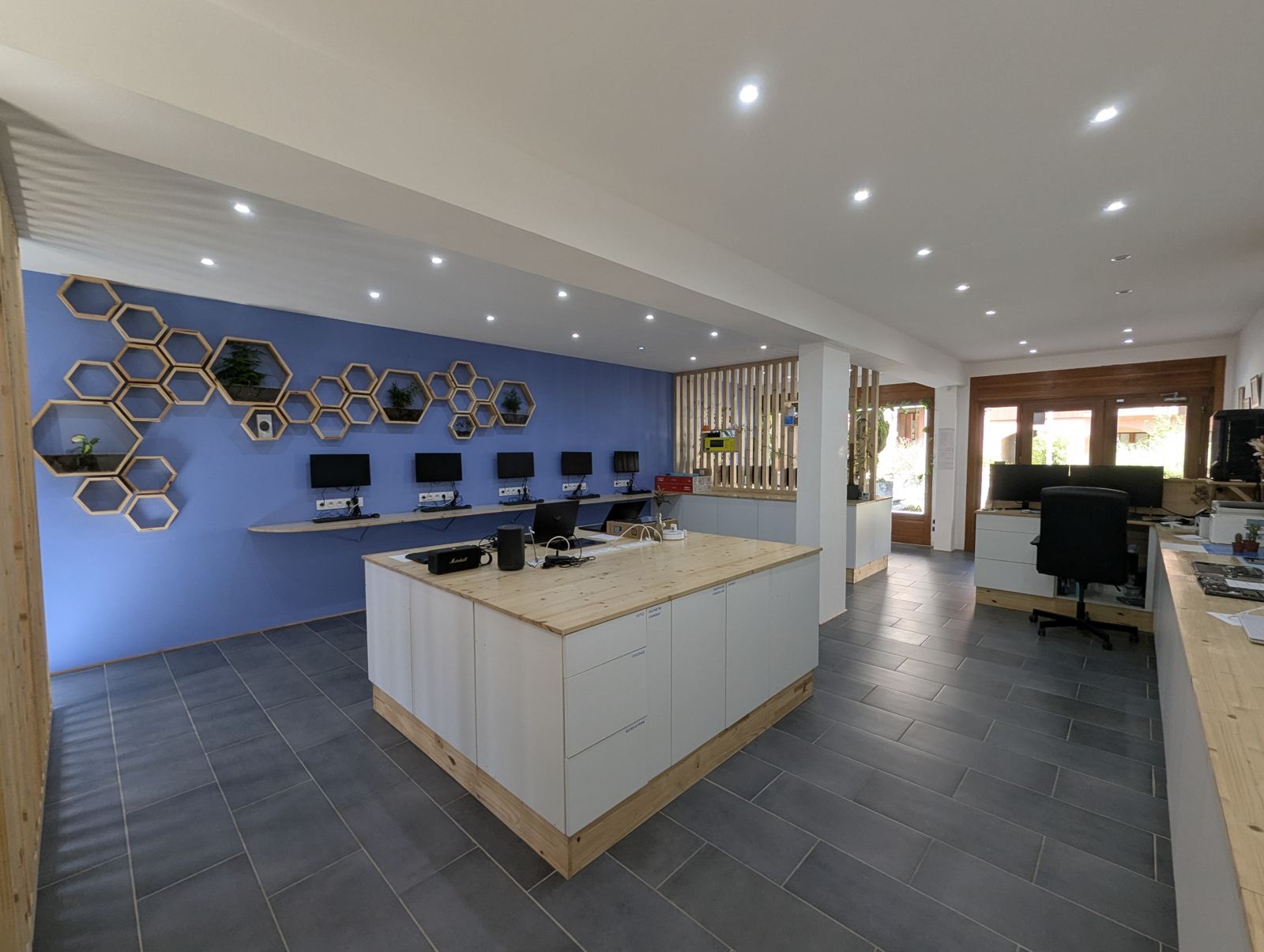 L'intérieur du magasin : un îlot central en bois et blanc, un mur bleu avec des étagères en nids d'abeille et des postes informatiques, un sol en grand carrelage gris.