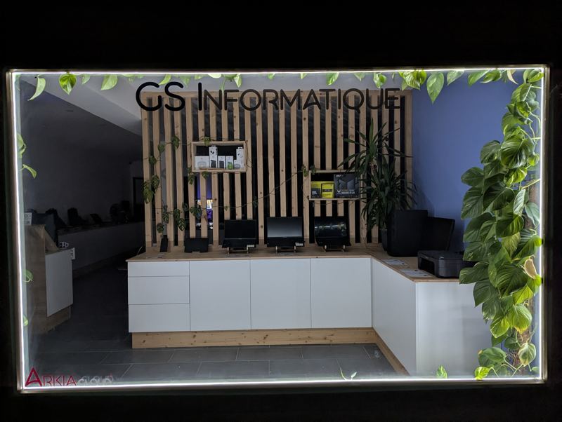 La vitrine du magasin éclairée le soir, encadrée de plantes, avec l'enseigne GS Informatique.